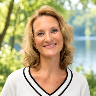 Headshot of Dena Koch, PMHNP-BC, smiling outdoors with shoulder-length wavy blonde hair and a white V-neck sweater with black trim.