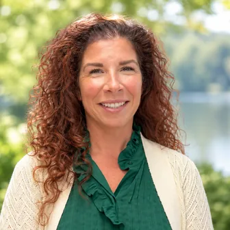 Headshot of Traci M. Williams, APRN, PMHNP-BC, smiling outdoors with long curly auburn hair, a green ruffled blouse and cream cardigan.