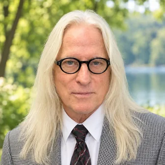 Headshot of Frank H. Deland, M.D., outdoors with long silver hair, round black glasses, a grey houndstooth jacket and dark tie.