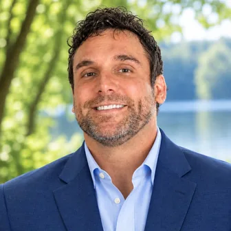 Headshot of Charles R. Noplis II, M.D., smiling outdoors with short dark curly hair and a beard, in a navy blazer and light blue shirt.
